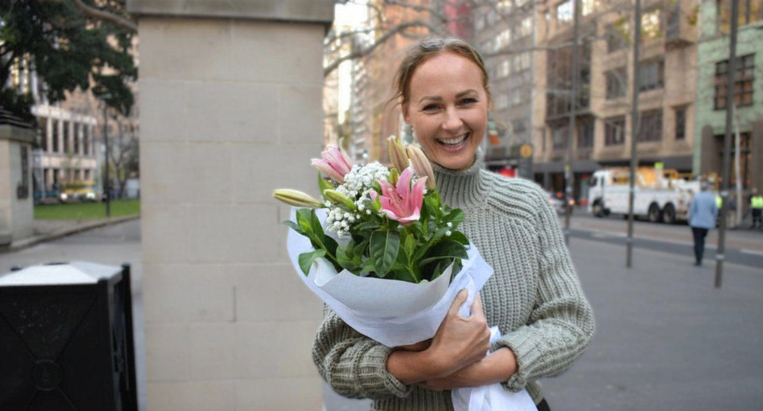 A smiling woman in a green knitted jumper holds a bouquet of pink lilies and white gypsophila on a city street.